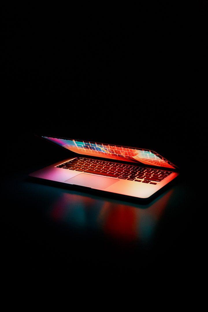 An almost closed laptop lit with bright colour against a black background